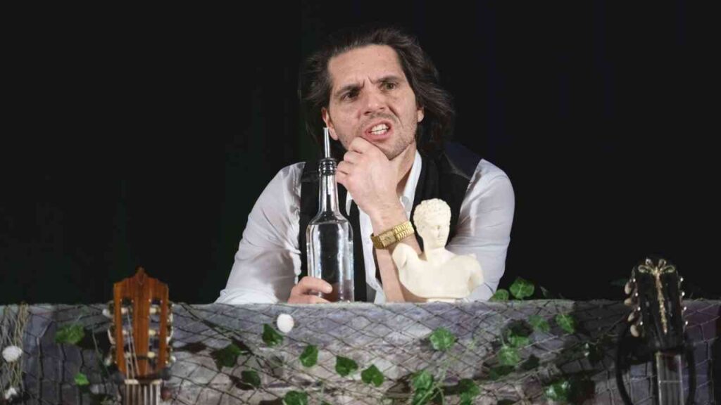 Ein Mann mit langen Haaren sitzt an einem Tisch mit einer Wasserflasche und einer Statue, umgeben von Gitarren.