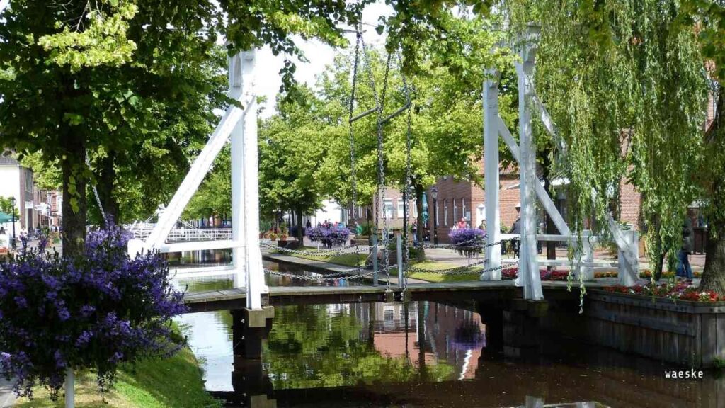Weiße Fußgängerbrücke über einen Kanal, umgeben von Bäumen und Blumen in Papenburg.