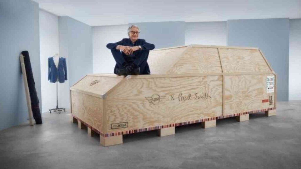 Paul Smith sitzt auf einem großen Holzkasten mit seinem Namen, umgeben von einem Anzug und einer minimalistischen Umgebung.