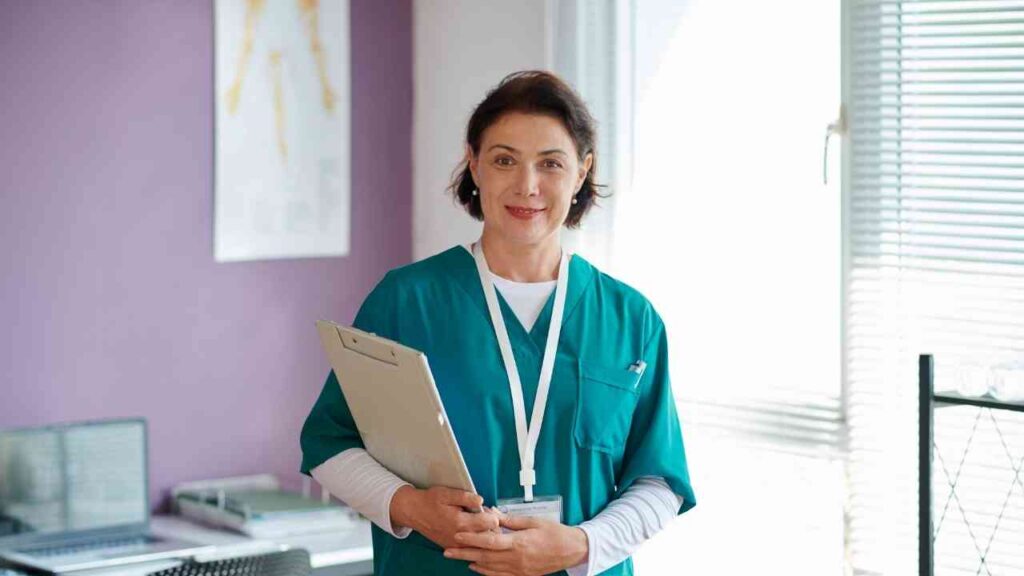 Frau in grüner Pflegekleidung mit Klemmbrett in einem Raum mit Fenster und natürlichem Licht.