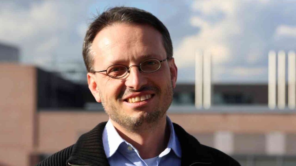 Professor mit Brille und lächelndem Gesicht, im Freien vor einem modernen Gebäude.