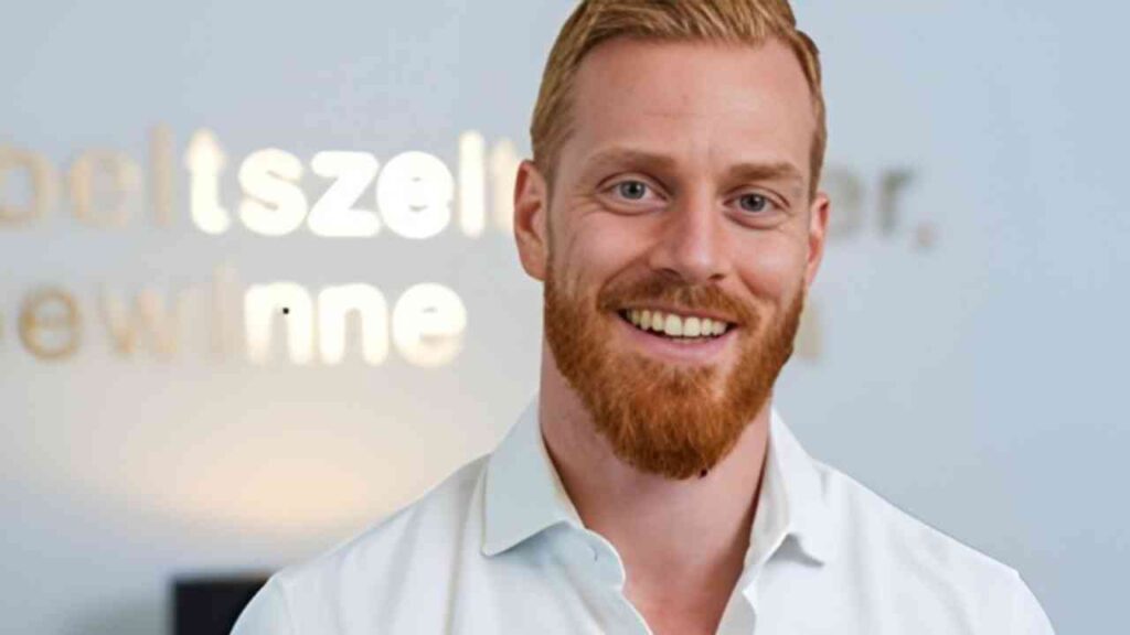 Lächelnder Mann mit rotem Bart und blonden Haaren, der ein weißes Hemd trägt und vor einem hellen Hintergrund steht.