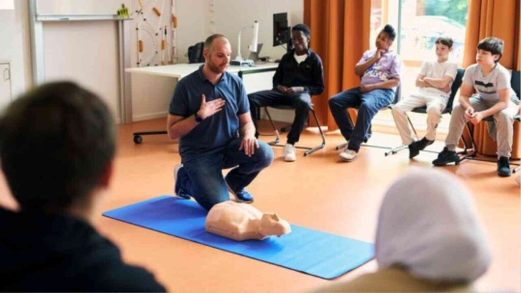 Ein Ausbilder demonstriert Wiederbelebungstechniken mit einer Übungspuppe vor einer Gruppe von Schülern.