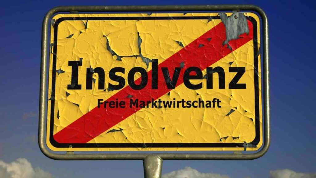 Ortsschild mit dem Wort „Insolvenz“ und durchgestrichenem Schriftzug „Freie Marktwirtschaft“ vor blauem Himmel – Symbolbild für steigende Insolvenzen