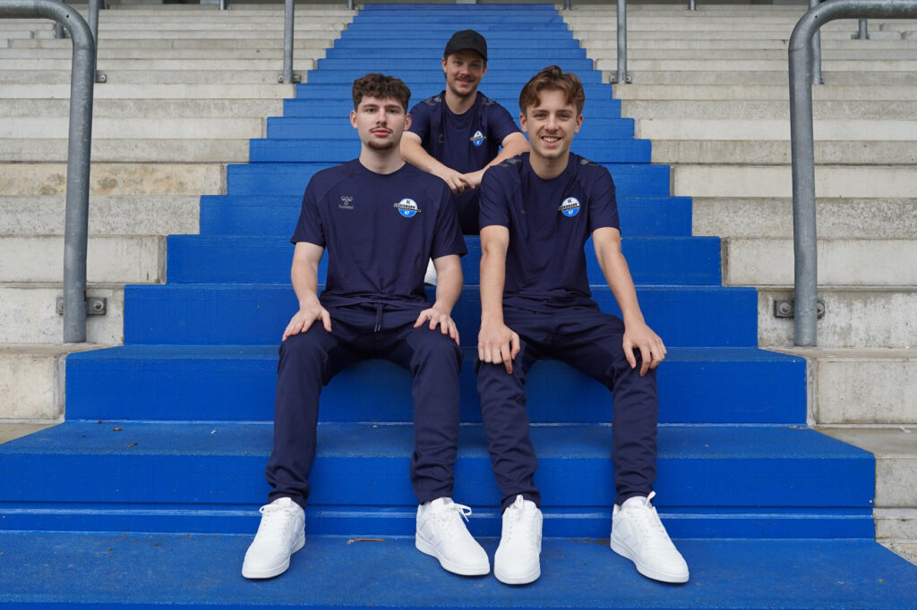 Drei Personen in sportlicher Kleidung sitzen auf blauen Treppenstufen in einem Stadion.