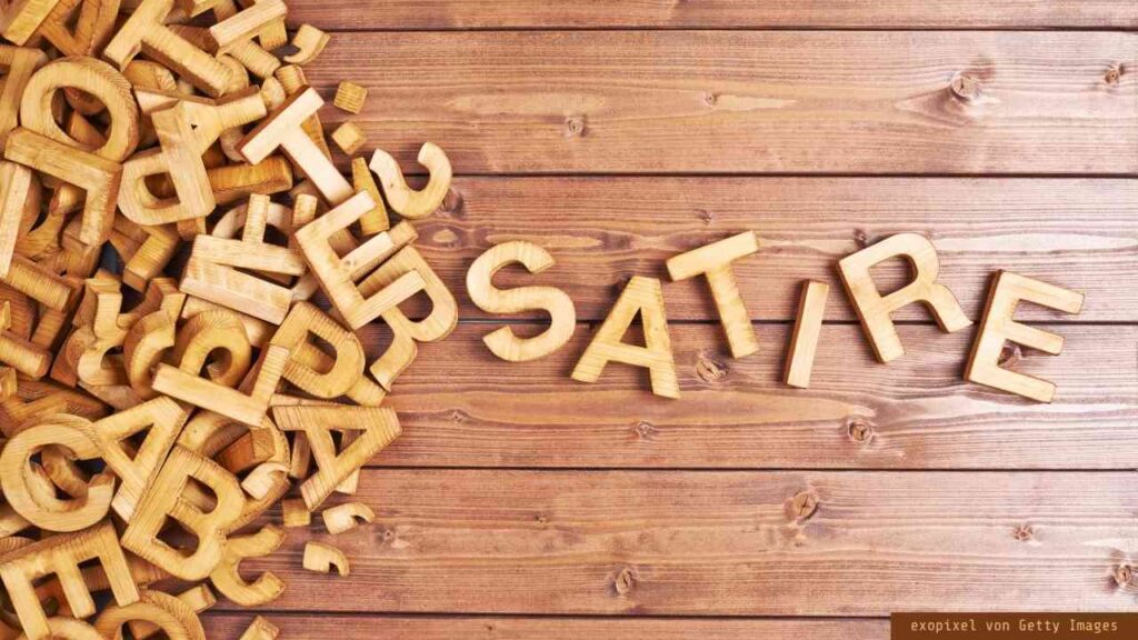 Holzbuchstaben auf einem Tisch, die das Wort 'SATIRE' bilden, umgeben von weiteren Buchstaben.