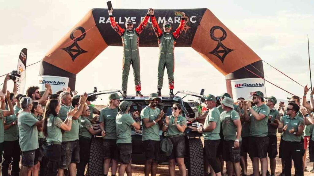 Zwei Rennfahrer stehen auf einem Fahrzeug und feiern ihren Sieg beim Rallye-Event, umgeben von jubelnden Teammitgliedern.