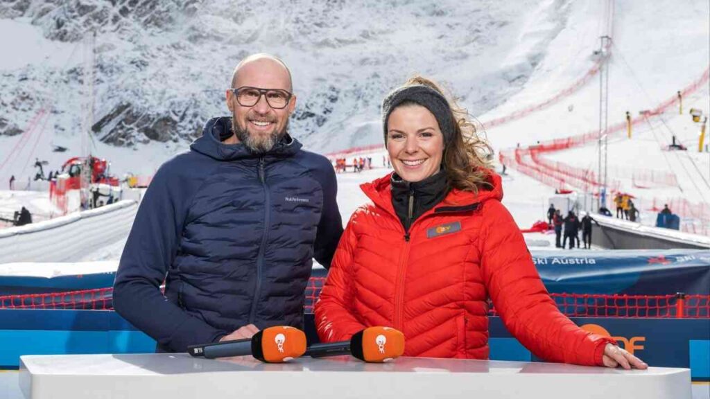 Zwei Moderatoren im Winteroutfit stehen vor einer Skipiste mit Schnee und Skiausrüstung im Hintergrund.
