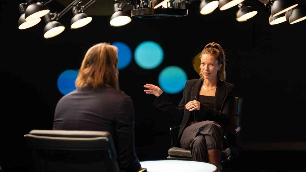 Zwei Personen in einem Interviewsetting mit Scheinwerfern, eine Frau spricht und gestikuliert.