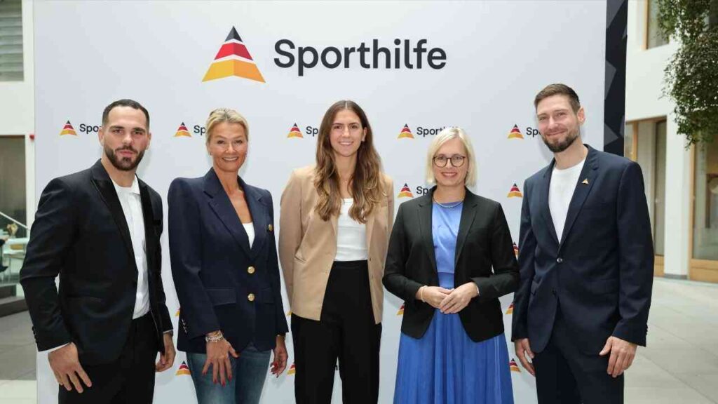 Fünf Personen stehen vor einem Sporthilfe-Logo, darunter Athleten und Vertreter, in einem modernen Innenraum.