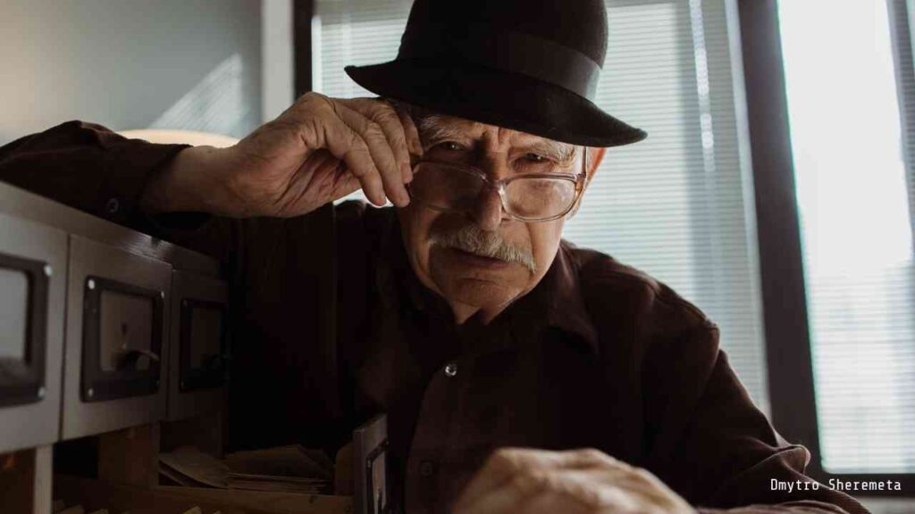 Älterer Mann mit Brille und Hut, der in einer Schublade mit Karteikarten schaut.