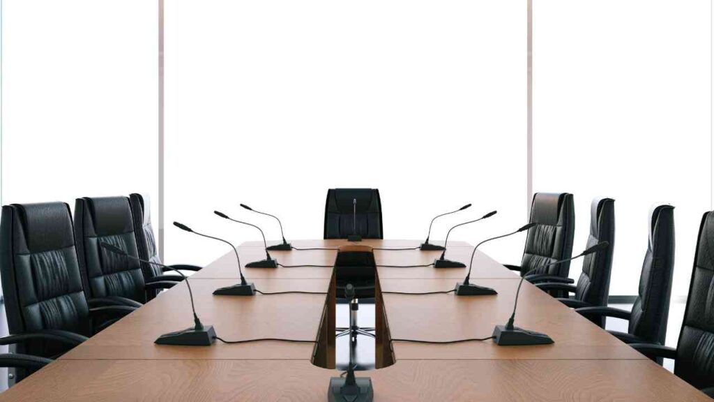 Konferenzraum mit einem langen Tisch und mehreren Mikrofonen sowie schwarzen Bürostühlen.