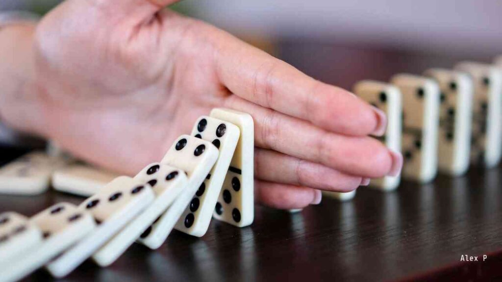 Eine Hand stoppt fallende Dominosteine auf einem Tisch, die in einer Reihe angeordnet sind.