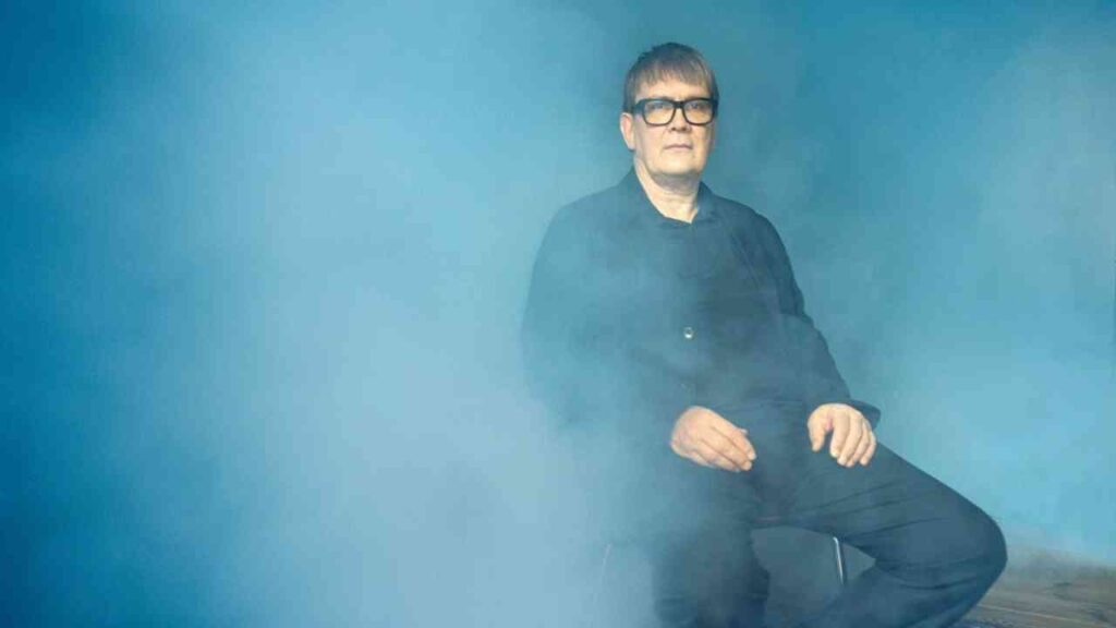 Mann mit Brille sitzt in einem blauen Nebel, trägt schwarze Kleidung und hat einen neutralen Gesichtsausdruck.