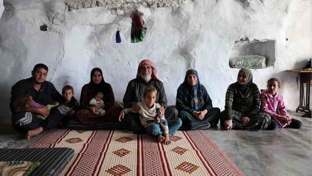 Eine Gruppe von sieben Personen in traditioneller Kleidung sitzt in einem Raum mit grauen Wänden und einem Teppich.