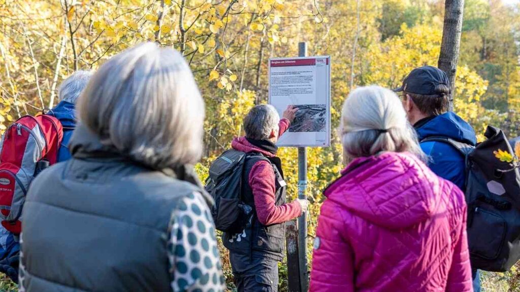 Gruppe von Wanderern betrachtet ein Informationsschild im Wald mit einer Karte und Hinweisen zu Wanderwegen.