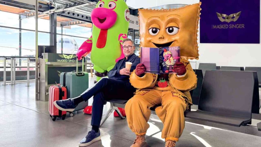 Mann in einem Wartebereich mit einer Tasse Kaffee, neben zwei maskierten Charakteren in bunten Kostümen.