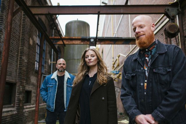 Drei Personen stehen in einer urbanen Umgebung, zwei Männer und eine Frau, mit unterschiedlichen Frisuren und Kleidungsstilen.