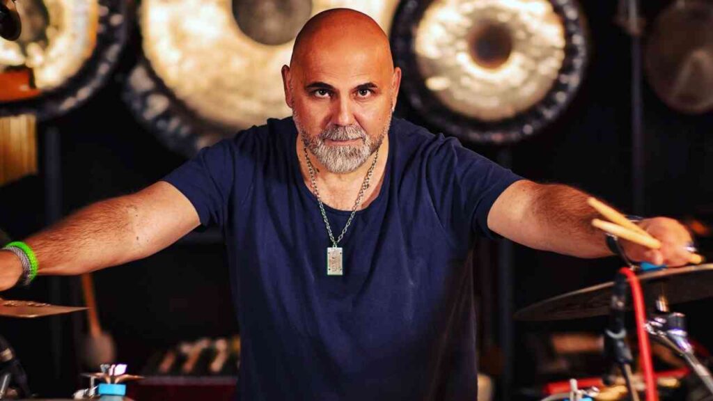 Mann mit Glatze und Bart, der an einem Schlagzeug mit Zimbeln steht und Stöcke in der Hand hält.