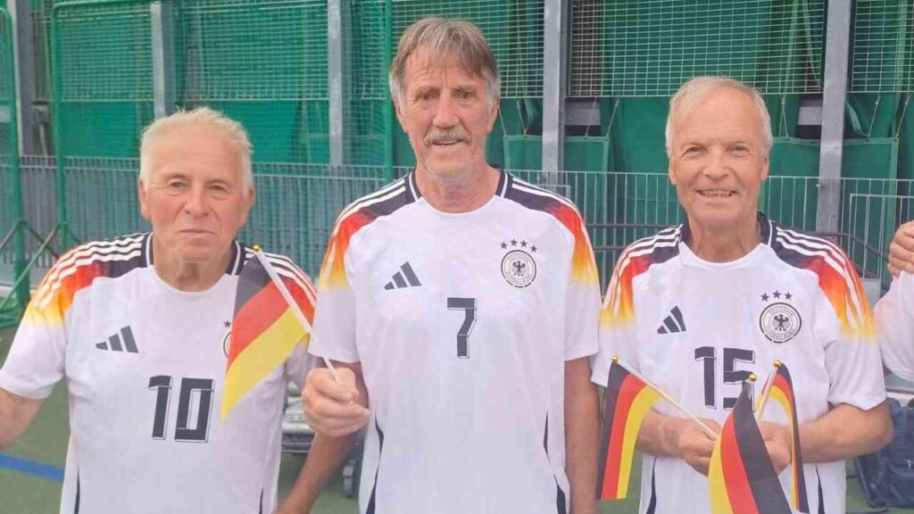 Drei ältere Männer in Fußballtrikots mit deutschen Flaggen und Nummern auf dem Trikot, lächelnd auf einem Sportplatz.