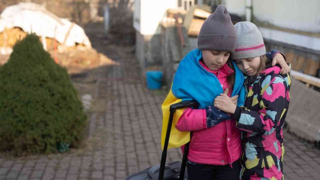 Zwei Kinder mit einem Koffer, eines trägt eine ukrainische Flagge, beide umarmen sich in einem Außenbereich.