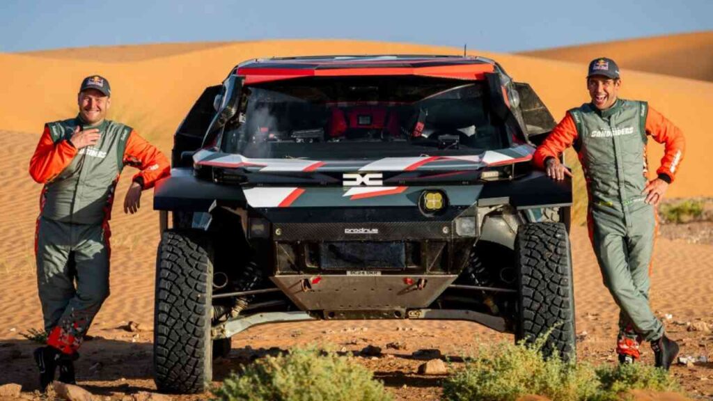 Zwei Fahrer in Rennanzügen stehen neben einem Dacia Sandrider in einer Wüstenlandschaft.