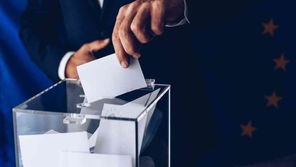 Eine Hand, die einen Stimmzettel in eine transparente Wahlurne einwirft, im Hintergrund eine europäische Flagge.