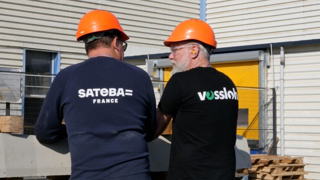 Zwei Bauarbeiter in orangefarbenen Helmen unterhalten sich vor einem Gebäude.