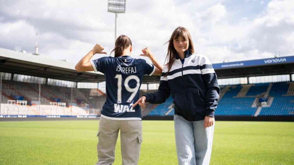 Zwei Frauen im Stadion, eine zeigt stolz auf das Trikot mit Nummer 19. Spielfeld im Hintergrund.