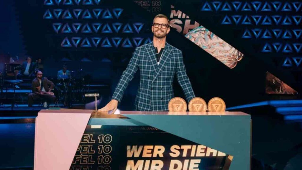 Joko Winterscheidt steht an einem Moderationstisch mit drei Auszeichnungen während einer Fernsehsendung.