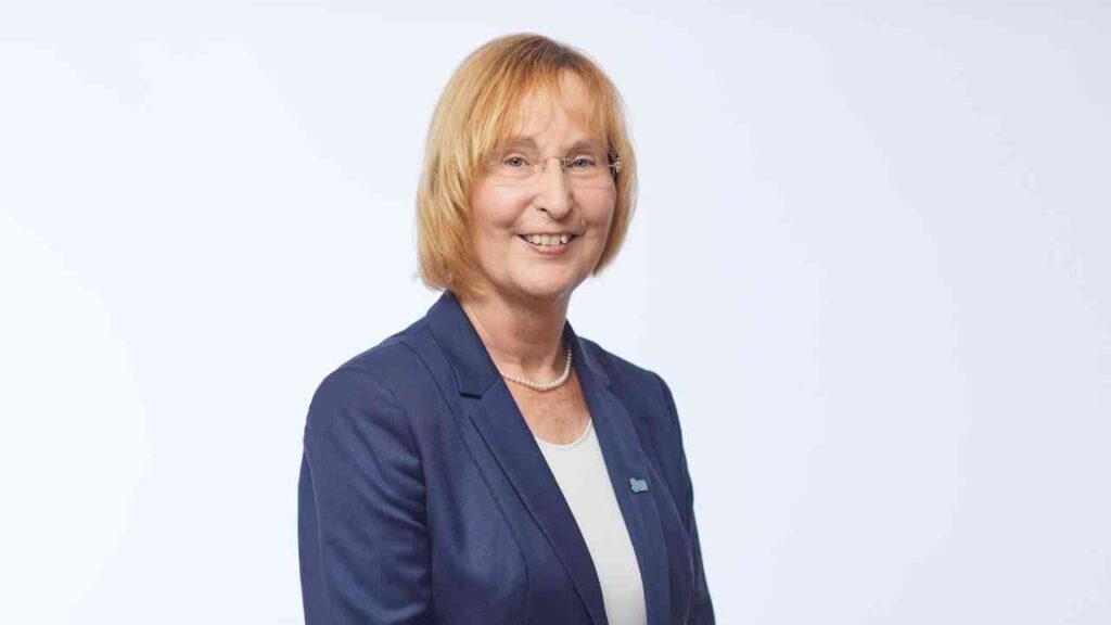 Frau mit blonden Haaren in einem blauen Blazer, lächelnd vor einem hellen Hintergrund.