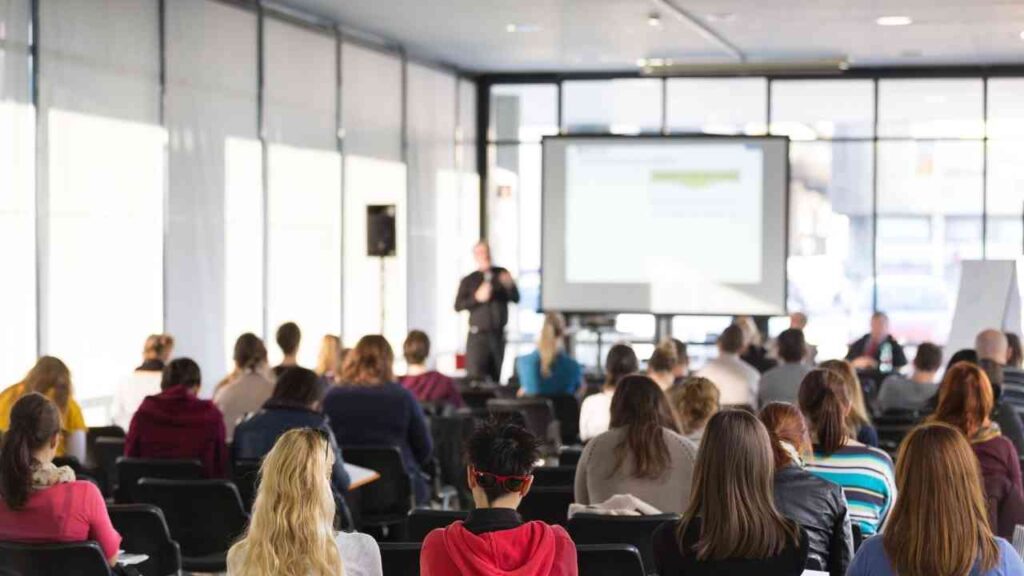 Mann präsentiert vor einer Gruppe von Teilnehmern in einem modernen Seminarraum mit Präsentationsbildschirm.