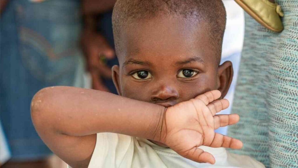 Ein kleines Kind mit brauner Haut und kurzen Haaren, das seine Hand vor dem Gesicht hält.