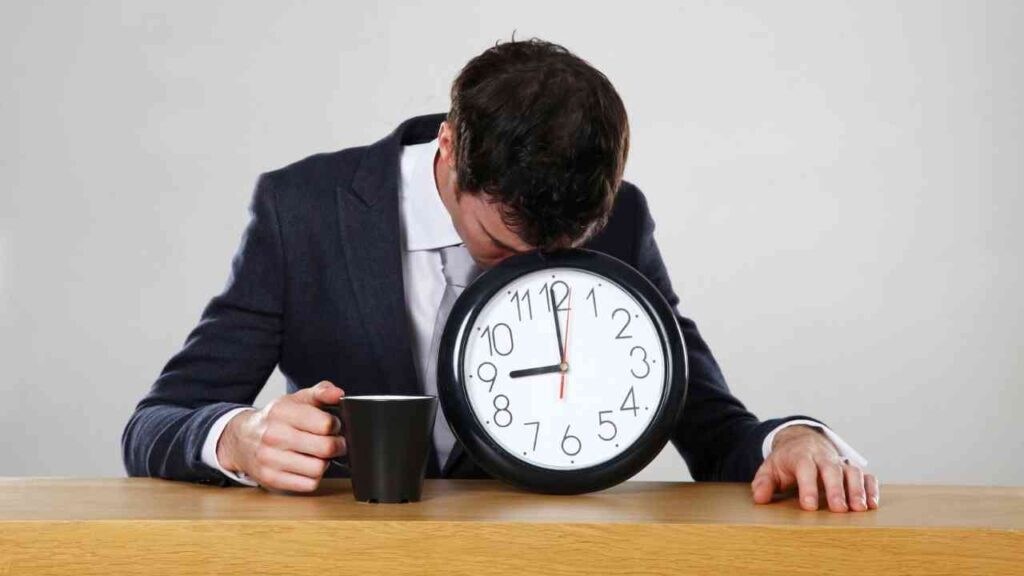 Mann im Anzug mit einer Kaffeetasse, der mit dem Kopf auf eine große Wanduhr lehnt.