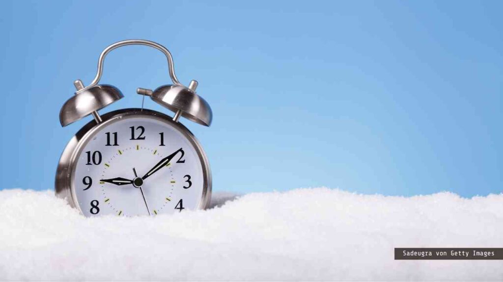 Wecker mit silbernem Gehäuse und Ziffernblatt, das in einer Schneedecke steht.