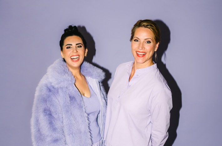 Zwei Frauen in lila Kleidung, eine trägt einen flauschigen Mantel, beide lächeln vor einem lila Hintergrund.