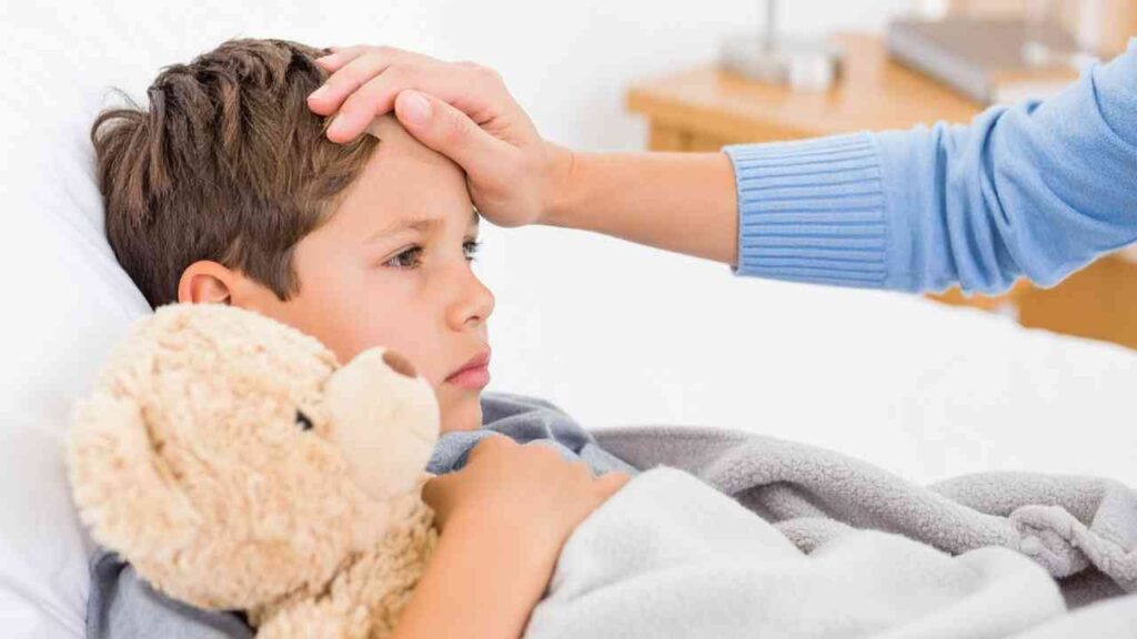 Ein Junge liegt im Bett mit einem Teddybär und wird von einer Person mit der Hand auf der Stirn berührt.