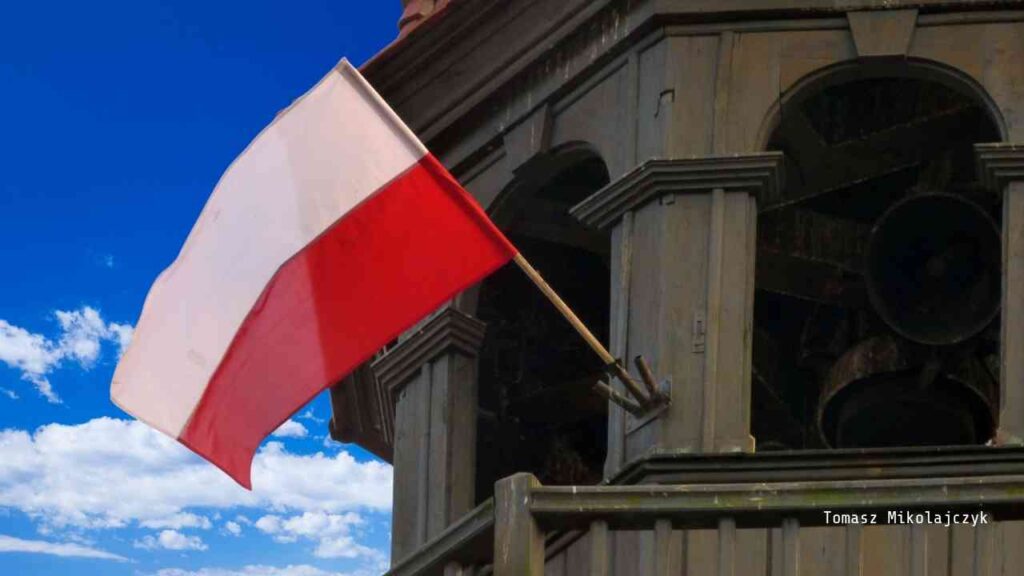 Polnische Flagge, die an einem Gebäude mit Glocken aufgehängt ist, vor einem blauen Himmel.
