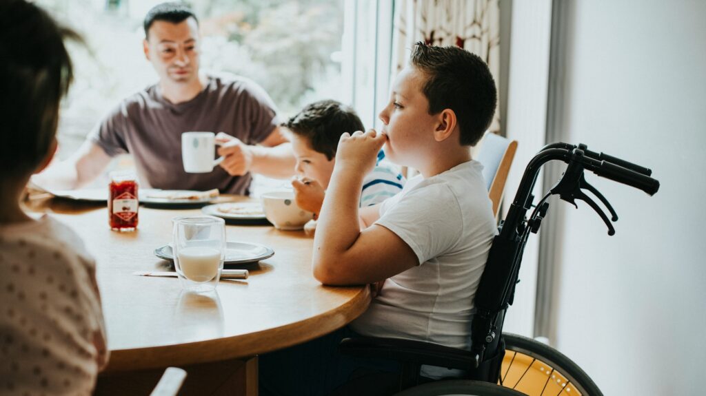 Junge im Rollstuhl sitzt am Tisch während einer Mahlzeit mit anderen Familienmitgliedern.