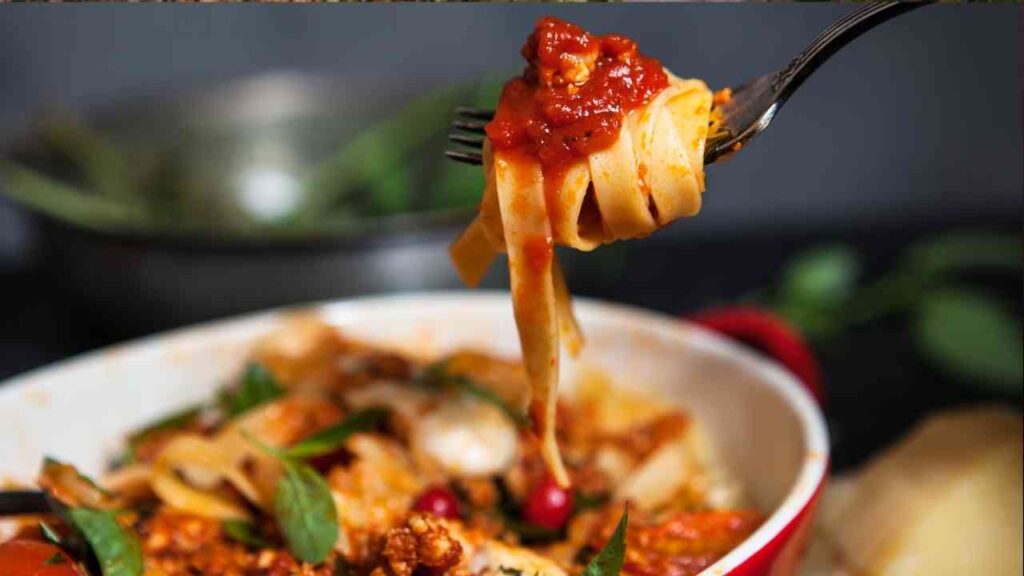 Eine Gabel mit Bandnudeln und Tomatensauce, die über einem Teller mit Pasta gehalten wird.