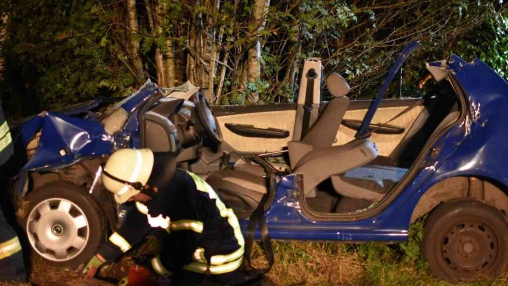 Verkehrsunfall in der Nacht: Feuerwehrmann rettet Person aus stark beschädigtem blauen Auto vor Bäumen.