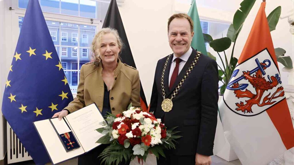 Ute Groth erhält das Bundesverdienstkreuz von einem Mann in offizieller Kleidung, umgeben von Flaggen.