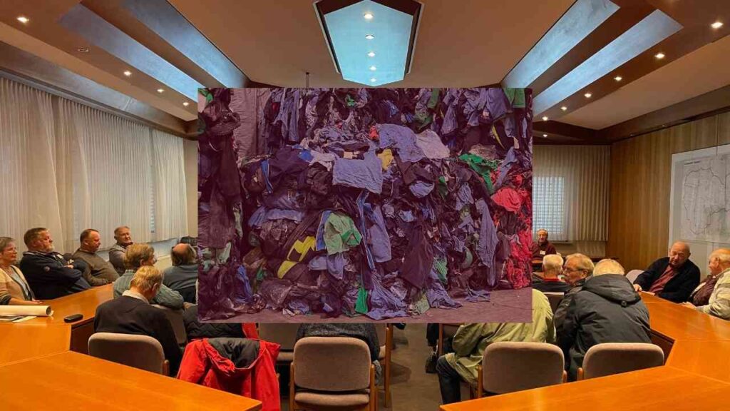 Bunte Stoffhaufen aus verschiedenen Kleidungsstücken in einem Besprechungsraum mit Teilnehmern im Hintergrund.