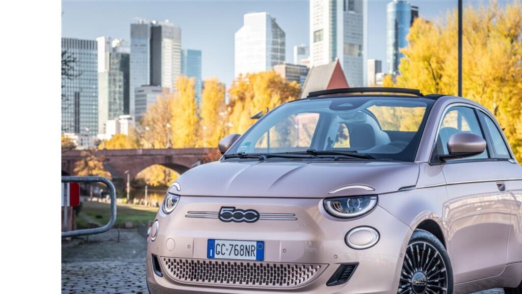 Rosa Fiat 500 Cabrio mit modernen Stadtgebäuden im Hintergrund und herbstlichen Bäumen.