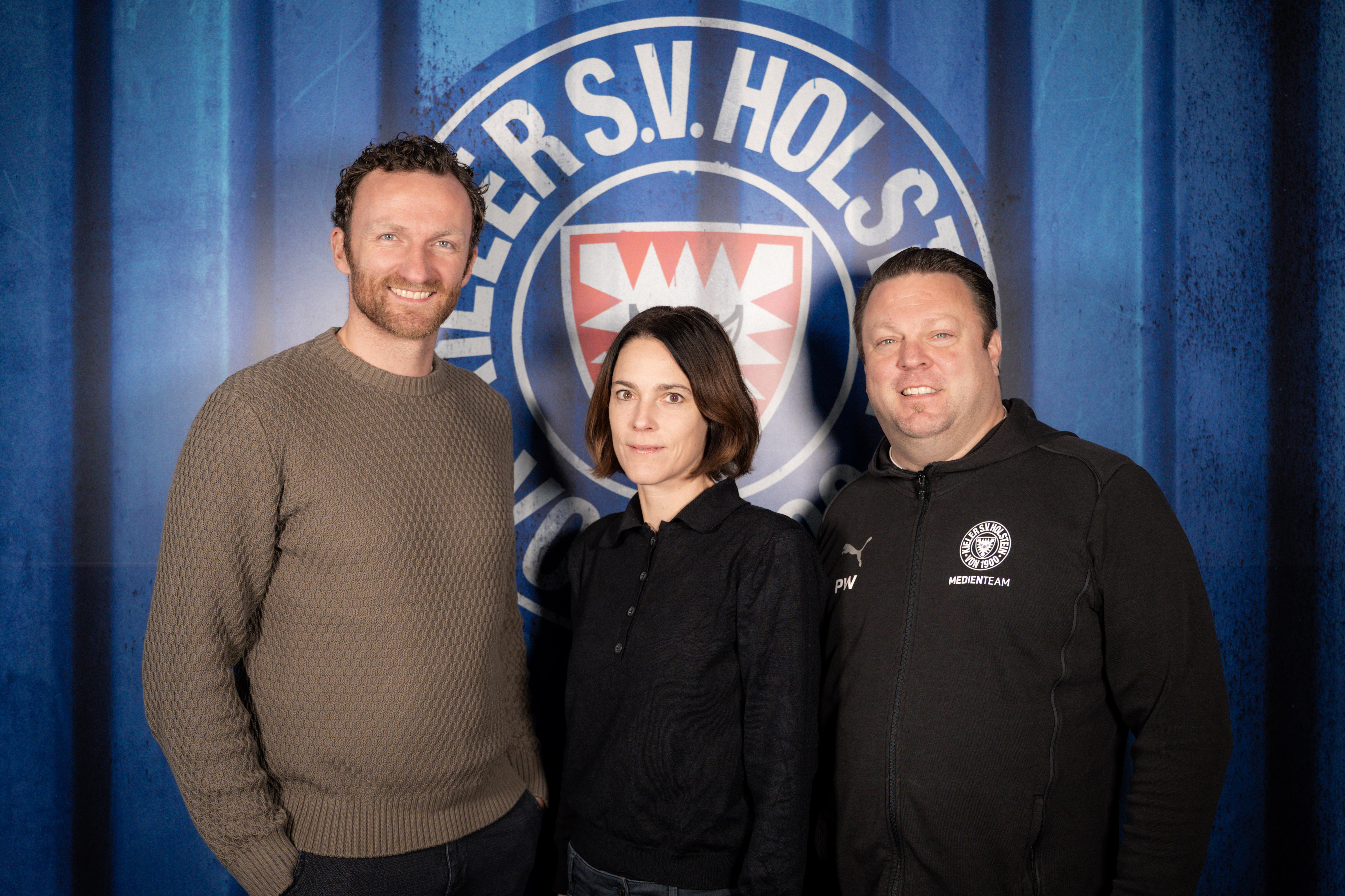 Drei Personen stehen vor einem blauen Hintergrund mit dem Logo von Holstein Kiel.