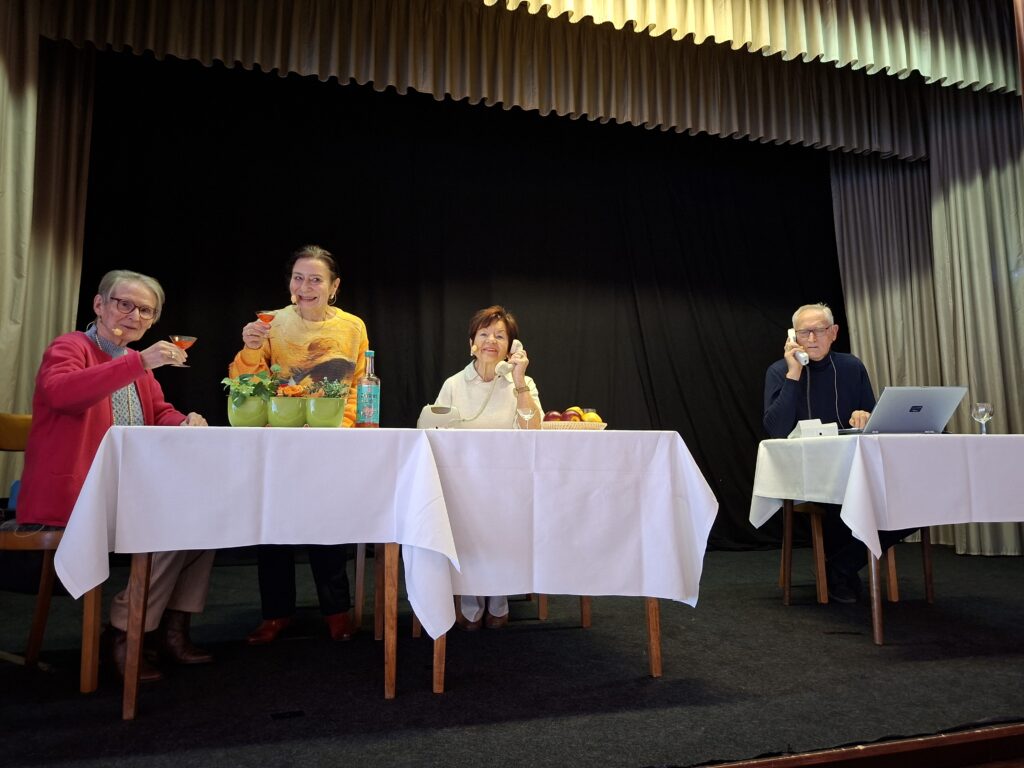 Vier Personen an einem Tisch, der mit weißen Tischtüchern und Obst dekoriert ist, während sie Getränke halten und miteinander kommunizieren.