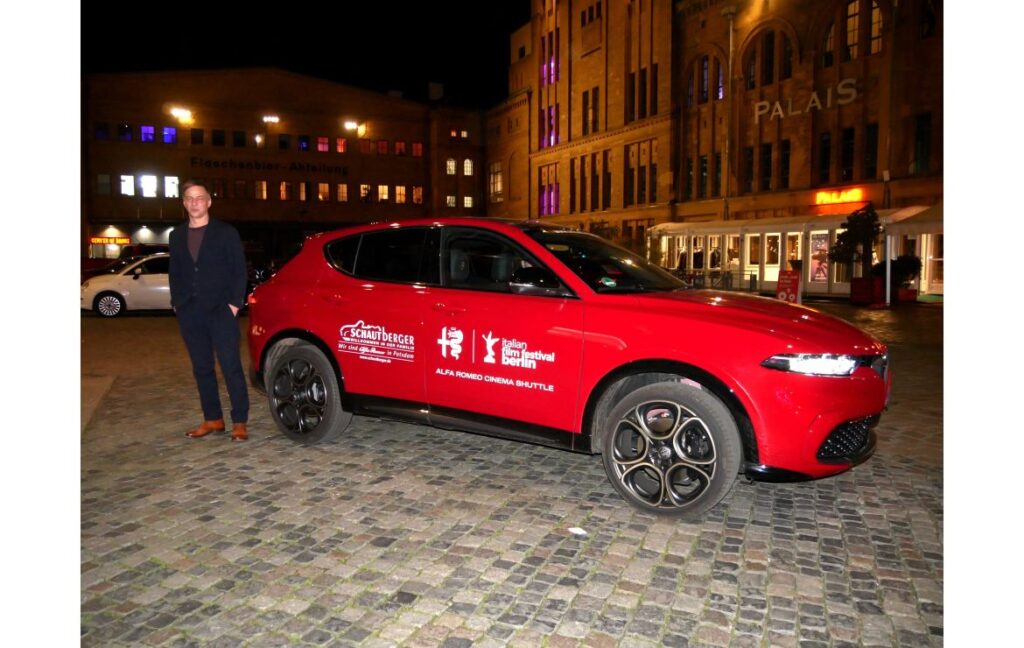 Rotes Auto mit Festival-Branding und einem Mann, der daneben steht, auf einem gepflasterten Platz.