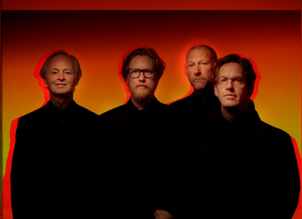 Vier Musiker des Helge Lien Trios in schwarzer Kleidung vor einem orange-roten Hintergrund.