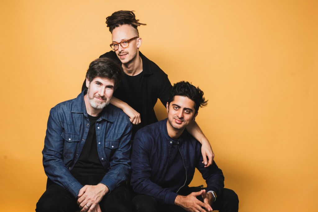 Philippe Lemm Trio mit drei Mitgliedern vor einem gelben Hintergrund, lächelnd und in lässiger Kleidung.