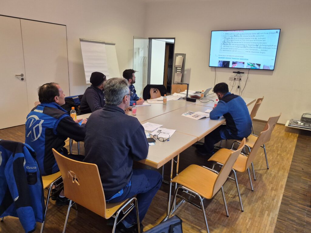 Gruppe von fünf Personen in einem Besprechungsraum, die an einem Tisch sitzen und auf einen Bildschirm schauen.